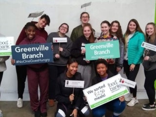 Students holding the signs of &quot;Food Bank&quot;, &quot;Greenville Branch #NoOneGoesHungry&quot;, &quot;We Packed 8,250 Meals&quot;, and such signs in front of #NoOneGoesHungry backdrop