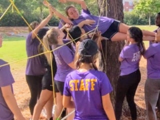 A student is being passed through a rope course by a group of students and being received by another group of students on the other side of the rope course